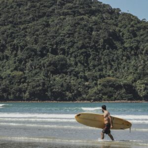 A surfer carrying his board on Borá beach, Brazil, with lush forest in the background.