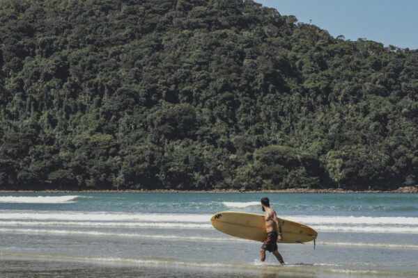 A surfer carrying his board on Borá beach, Brazil, with lush forest in the background.