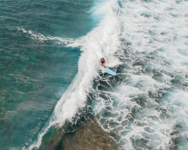 Drone shot capturing a surfer riding a wave in the clear blue waters of Hawaii.